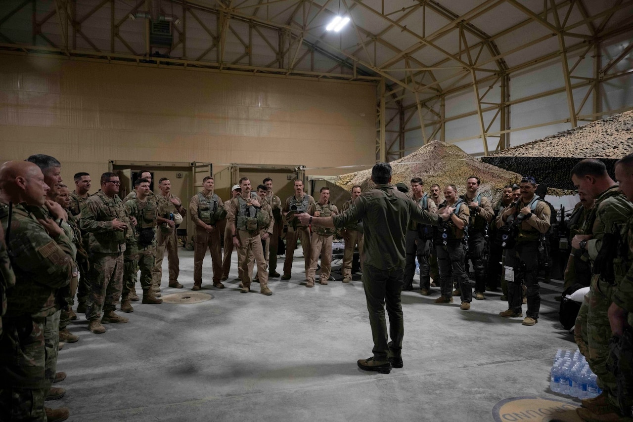 Dozens of people in camouflage military uniforms stand in a semicircle around a man in casual attire.