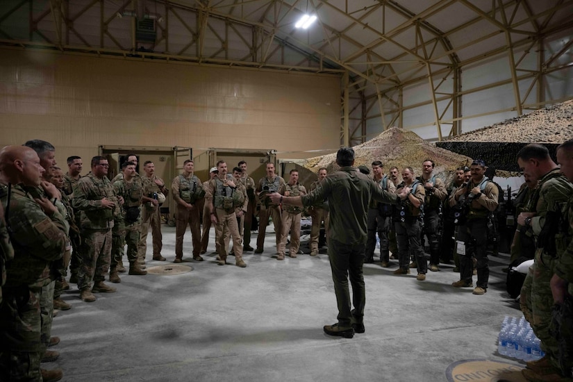 Dozens of people in camouflage military uniforms stand in a semicircle around a man in casual attire.