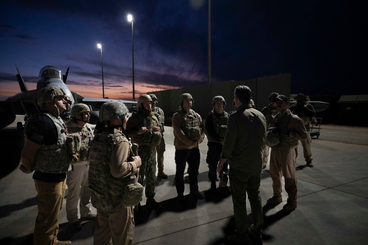A dozen people in camouflage military uniforms meet with a man in civilian attire near a helicopter, outside at night.