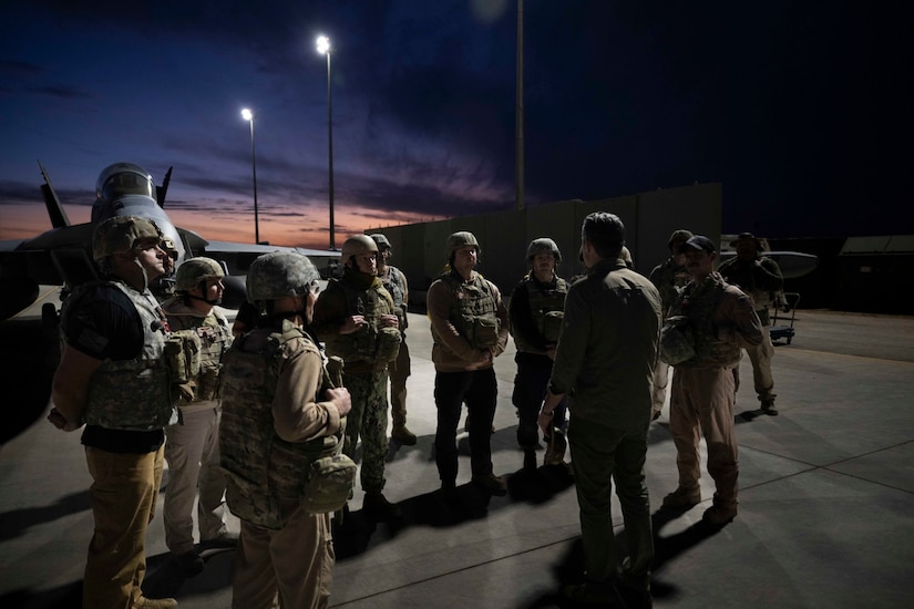 A dozen people in camouflage military uniforms meet with a man in civilian attire near a helicopter, outside at night.