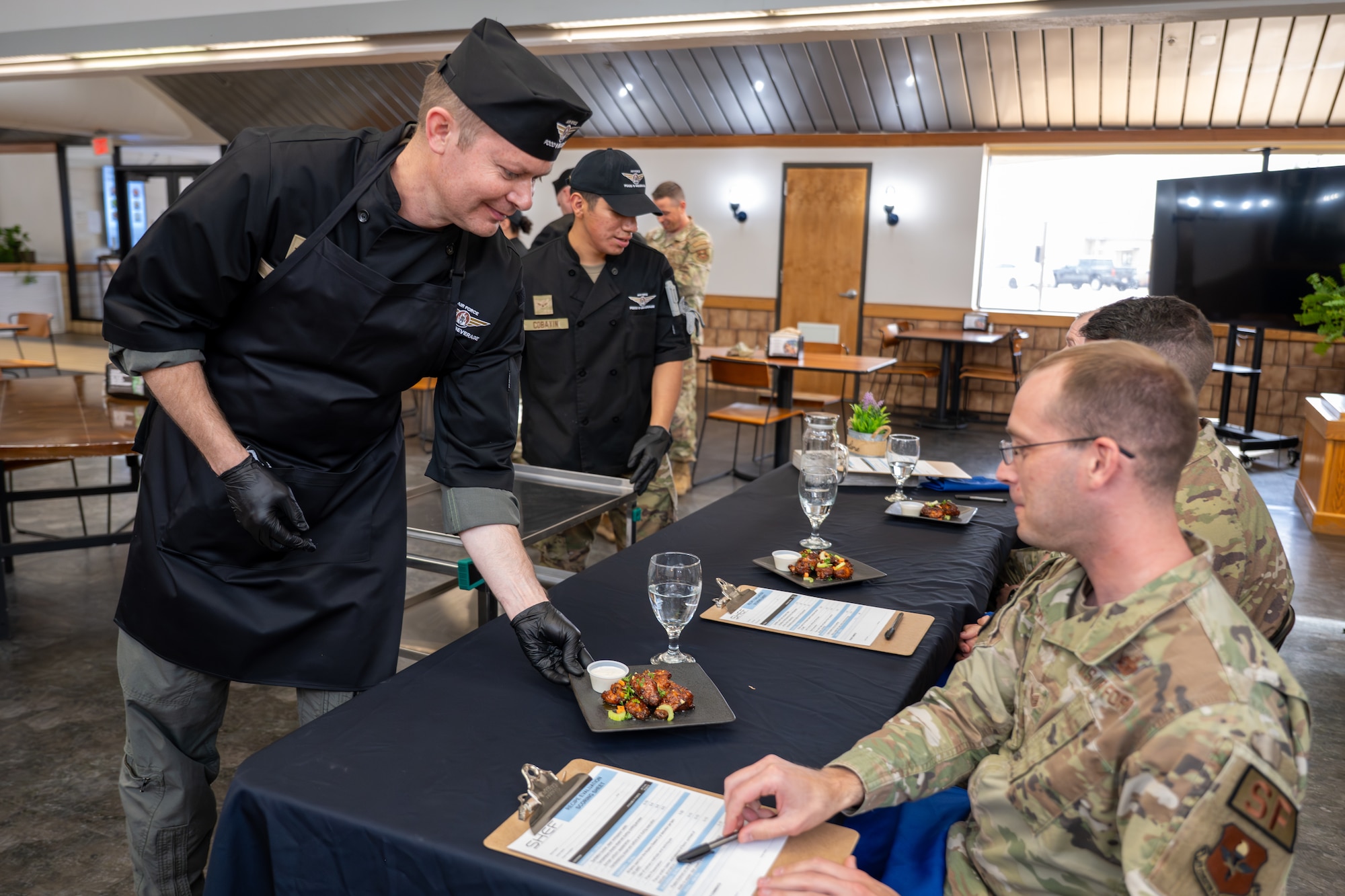 U.S. Air Force Col. Richard Kind, 97th Air Mobility Wing commander (left), and Airman 1st Class Jonathan Cobaxin, 97th Force Support Squadron dining facility shift worker (right), serve wings to the wing commander’s front office staff during an immersion tour competition hosted by the 97th Force Support Squadron at Altus Air Force Base, Oklahoma, March 23, 2026. The pair won the competition based on the judges’ scores. (U.S. Air Force photo by Airman 1st Class Nathan Langston)