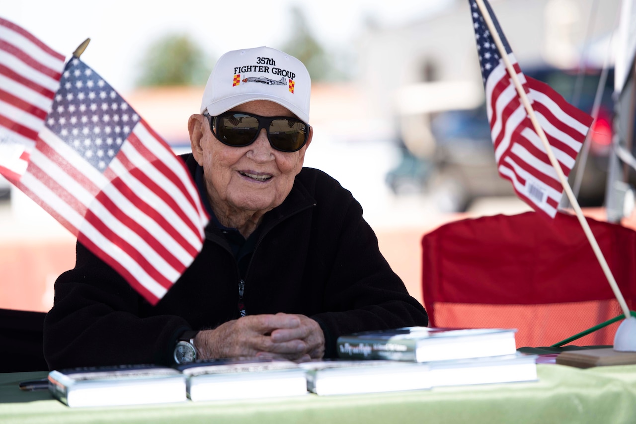 An elderly man wearing a hat and sunglasses sits at an outdoor table with books and small American flags displayed in front of him.