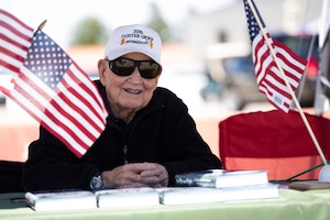 An elderly man wearing a hat and sunglasses sits at an outdoor table with books and small American flags displayed in front of him.