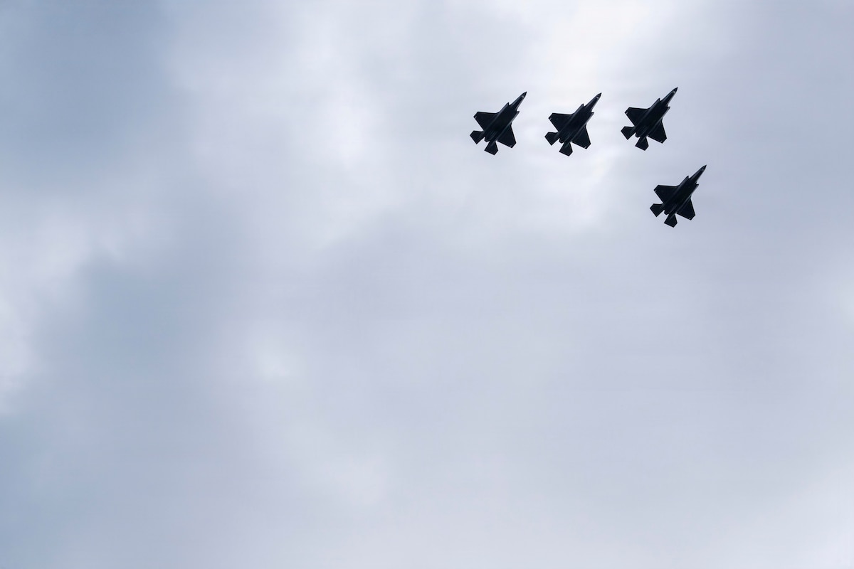 Four military fighter aircraft fly overhead.