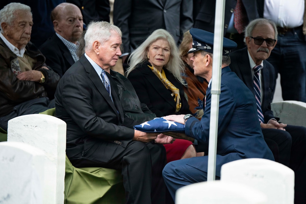 A man in a military dress uniform kneels to give a folded American flag to a man in a suit seated next to several other people in business attire.