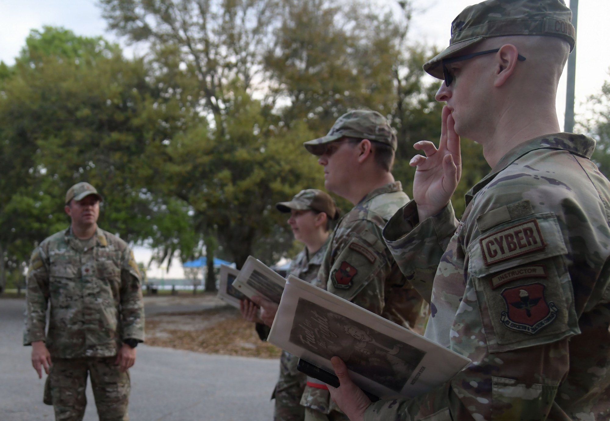 A man in military uniform speaks to others while holding a folder.