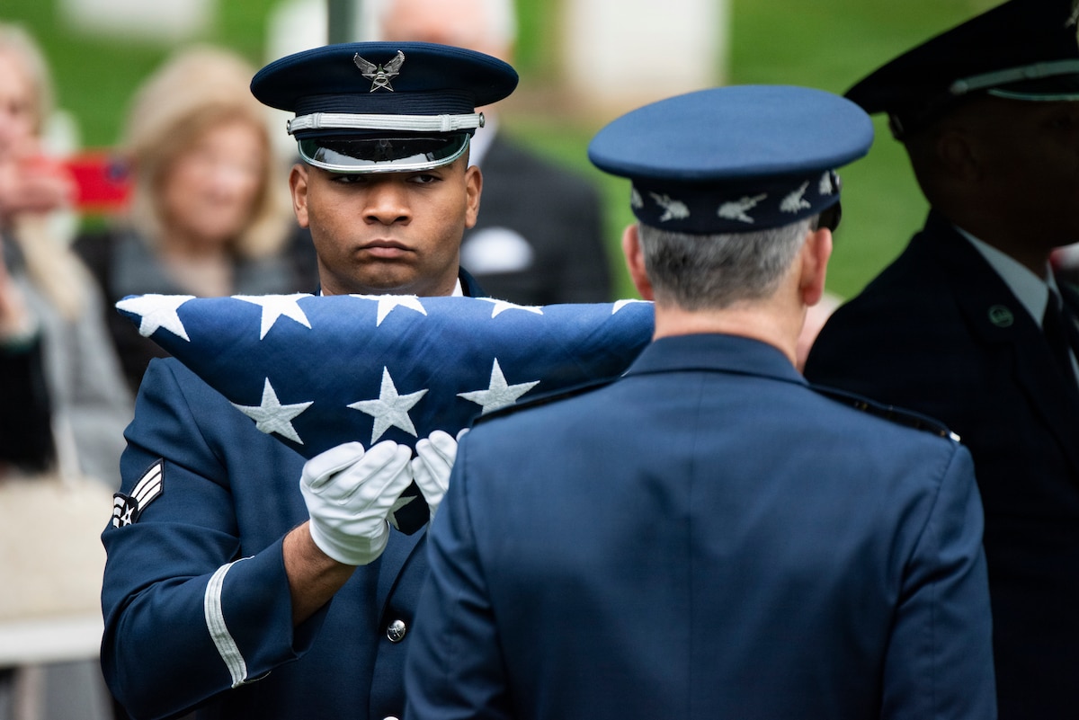 A man in a military dress uniform holds the American flag in front of another man in similar attire.