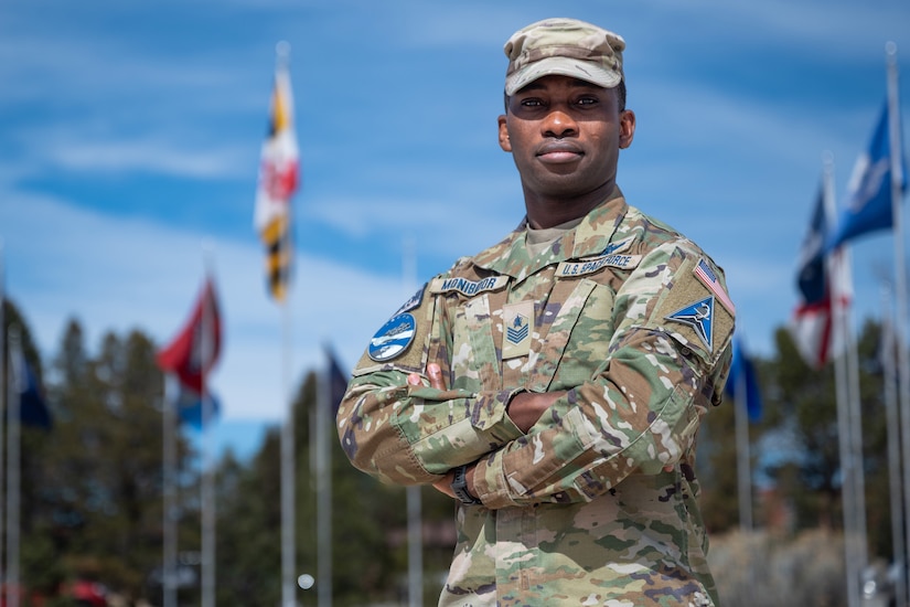 A guardian in a camouflage uniform stands with his arms folded outdoors during daytime, with blurry flags on poles and trees in the background.