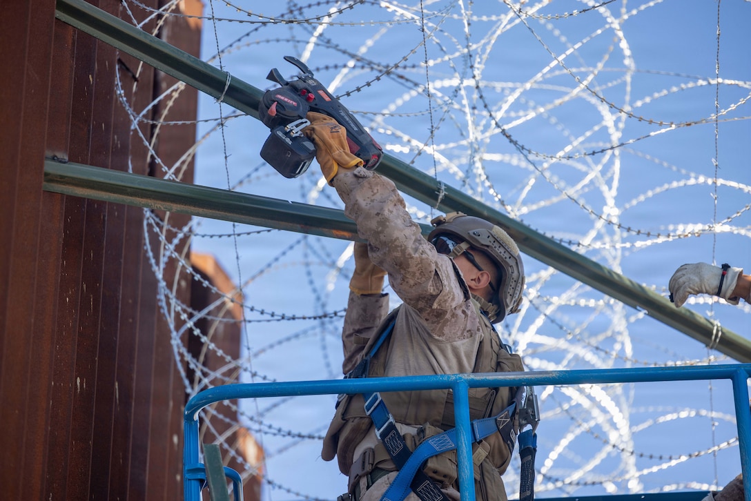 A Marine uses a tool to secure concertina wire overhead against a large, brown wall.