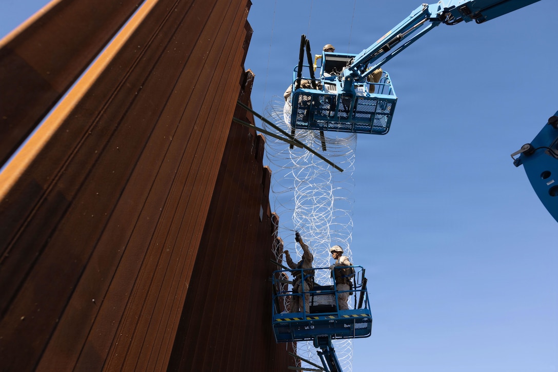 Four Marines , two in raised buckets, work with concertina wire against a large, high, dark brown fence.