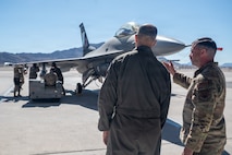 An Airmen points towards an F-16 Fighting Falcon aircraft while he gives a brief to Gen. Spain. The aircraft is static on the flightline with Airmen loading up weapons.