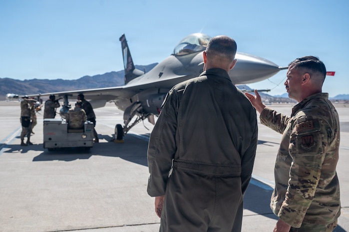 An Airmen points towards an F-16 Fighting Falcon aircraft while he gives a brief to Gen. Spain. The aircraft is static on the flightline with Airmen loading up weapons.
