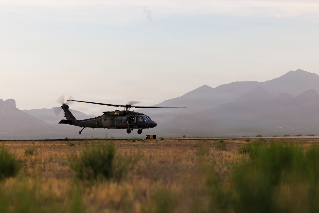 A helicopter flies low to the ground in a desert-like field with mountains in the distance.