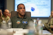 Gen. Adrian Spain sits at a conference table during a mission brief with Airmen sitting in the background, out of focus.