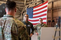 Gen. Adrian Spain and Chief Master Sgt. Jeremy Unterseher look at an Airman giving a brief inside a hangar, with a U.S.A flag in the background.