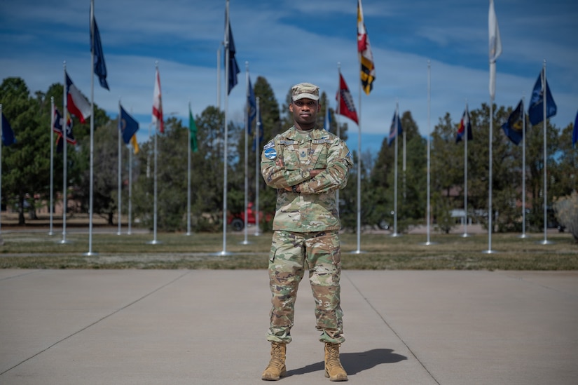 A guardian in a camouflage uniform stands with his arms folded outdoors during daytime, with flags on poles and trees in the background.