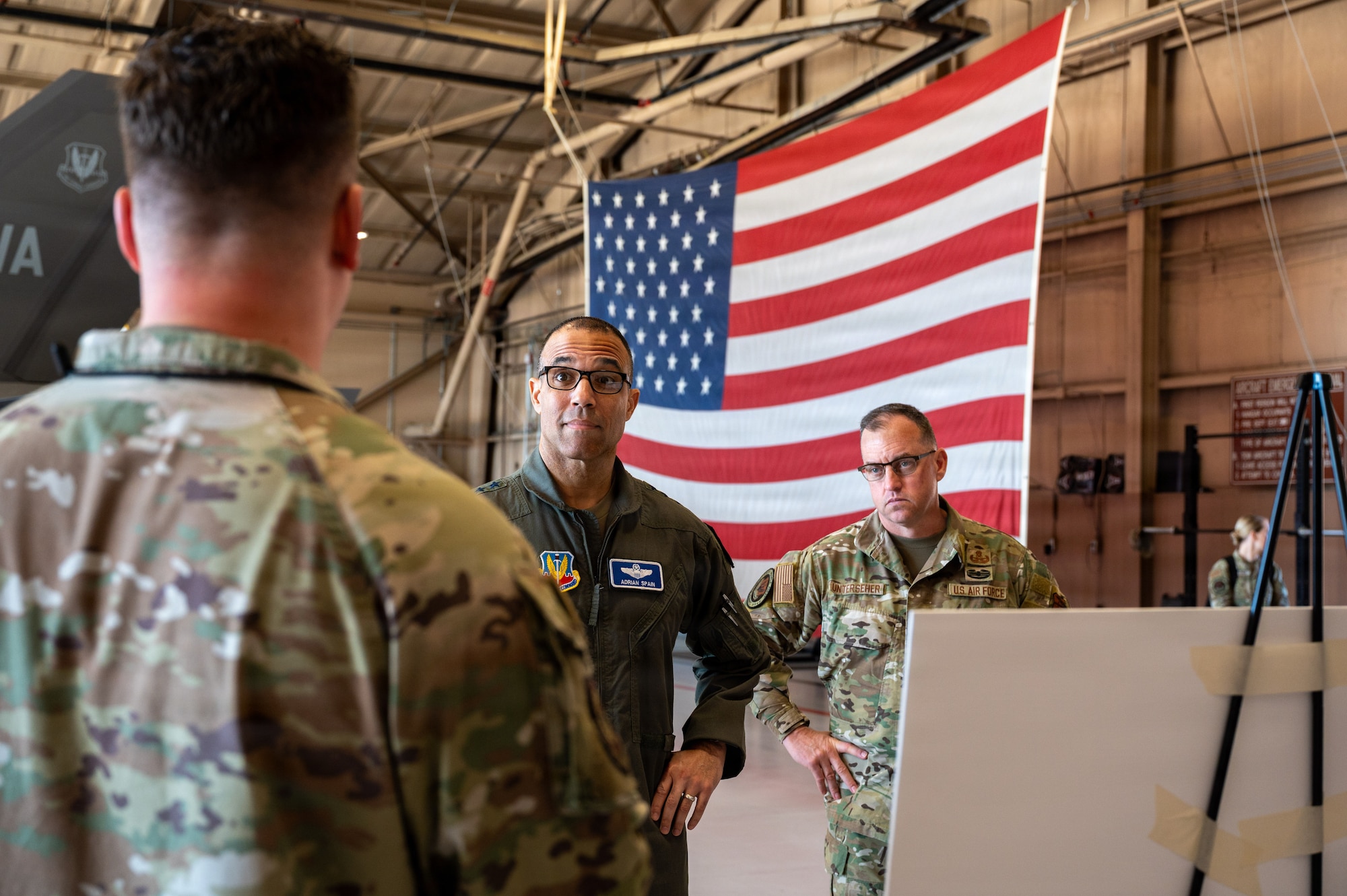 Gen. Adrian Spain and Chief Master Sgt. Jeremy Unterseher look at an Airman giving a brief inside a hangar, with a U.S.A flag in the background.