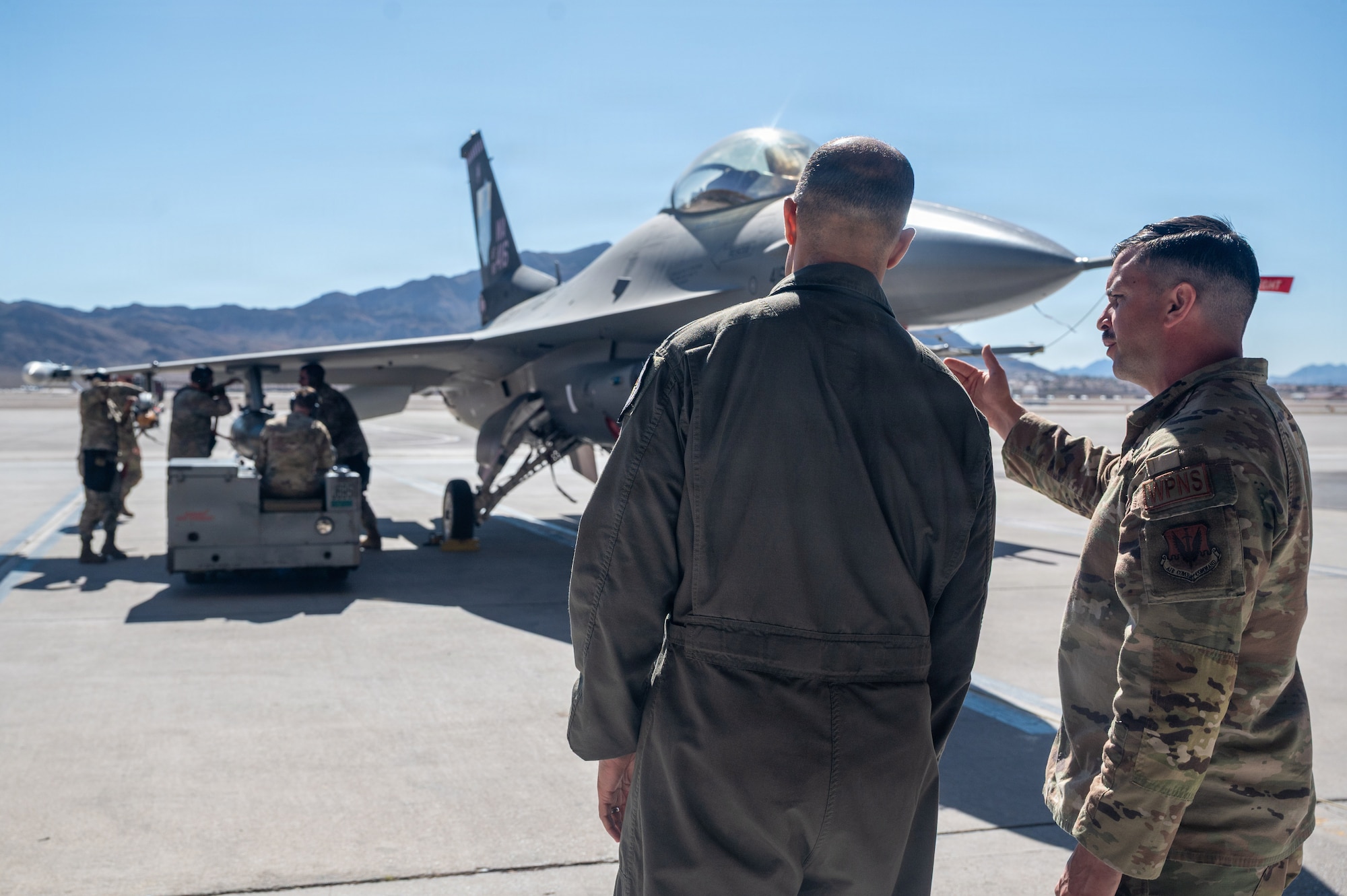 An Airmen points towards an F-16 Fighting Falcon aircraft while he gives a brief to Gen. Spain. The aircraft is static on the flightline with Airmen loading up weapons.