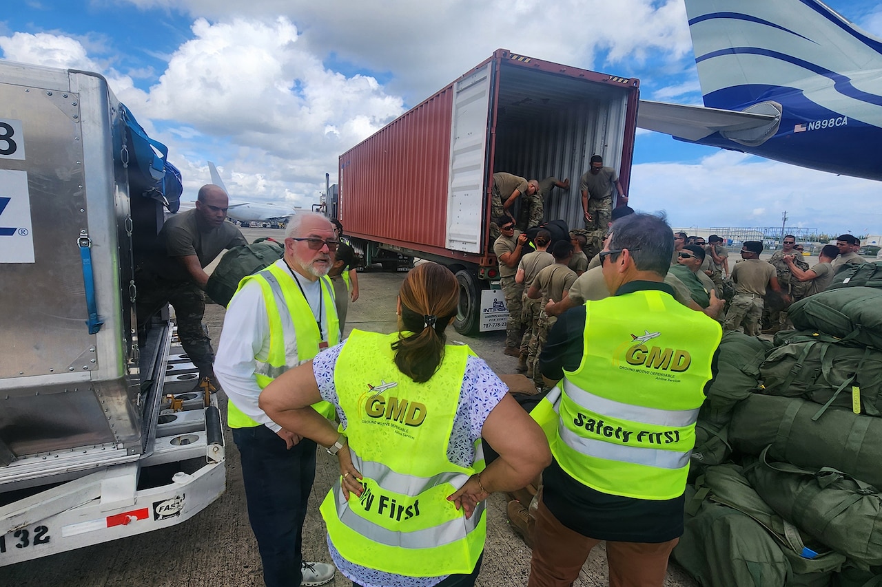 Three people wearing casual attire and reflective vests talk to each other while standing on a tarmac as dozens of people in camouflage military uniforms unload supplies from the back of a tractor trailer.