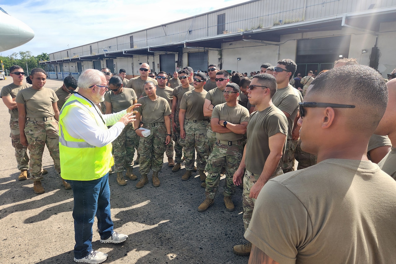 A man wearing casual attire and a reflective vest talks to a group of people in camouflage military uniforms standing outside on a tarmac with a building in the background.