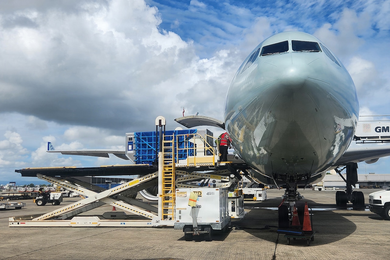 A person wearing a reflective vest uses a large machine to load equipment onto a plane parked on a tarmac.