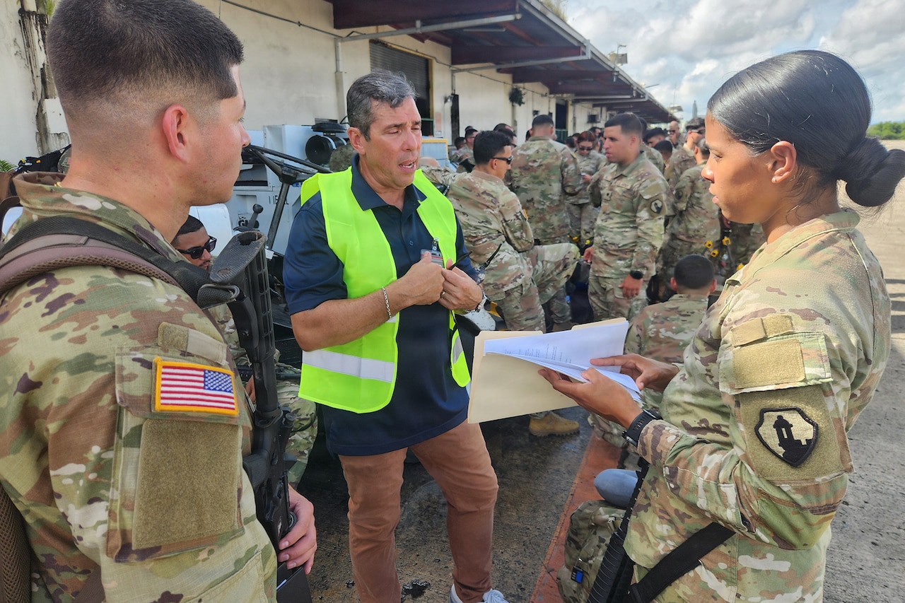 A man wearing business casual attire and a reflective vest talks to two people in camouflage military uniforms standing outside of a building on a tarmac; there are several other people in camouflage military uniforms standing in the background.
