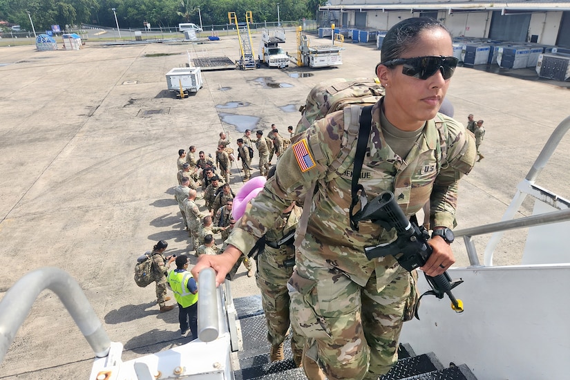 A woman wearing a camouflage military uniform, carrying a training rifle, walks up a flight of mobile stairs to an airplane as several people in similar attire follow behind her shaking hands with people on the tarmac.