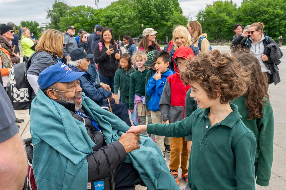 A veteran sitting in a wheelchair shakes hands with a child at an outdoor plaza where other kids, veterans and other citizens gather.