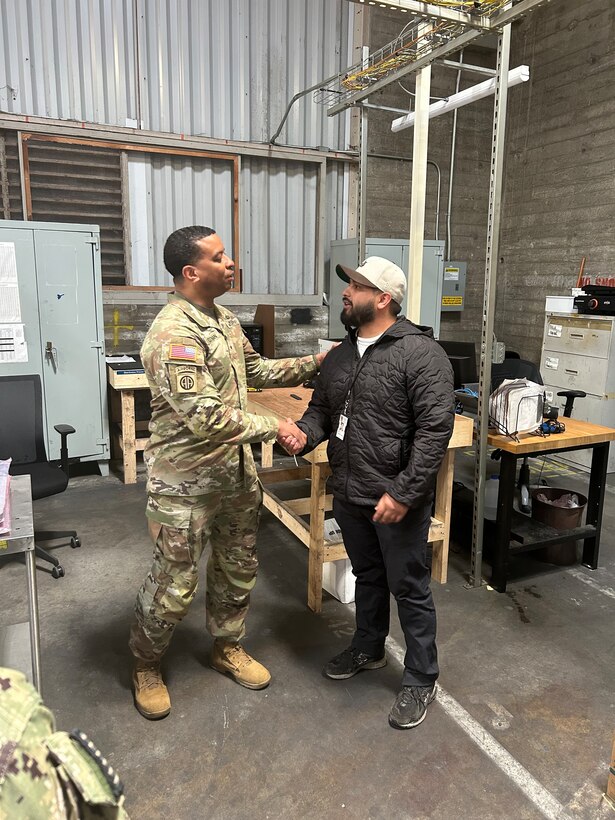 A military member shaking hands with a civilian employee in a warehouse environment.