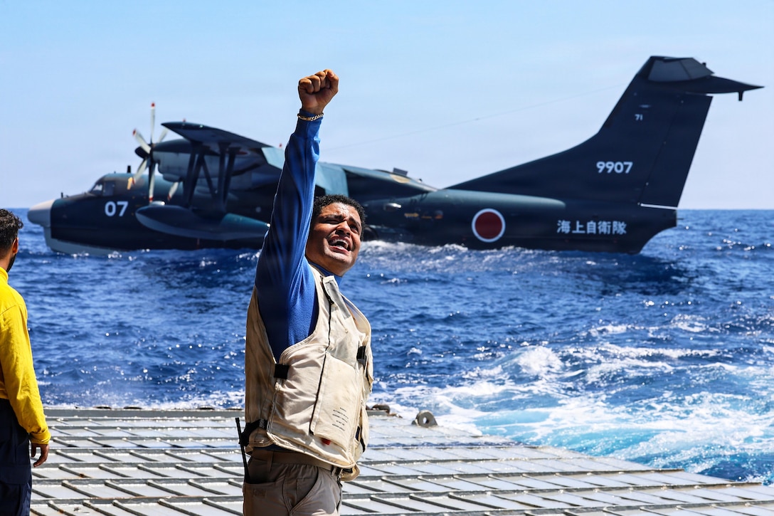 A sailor raises their fist while standing on a ship at sea as a black Japanese aircraft floats in the background.