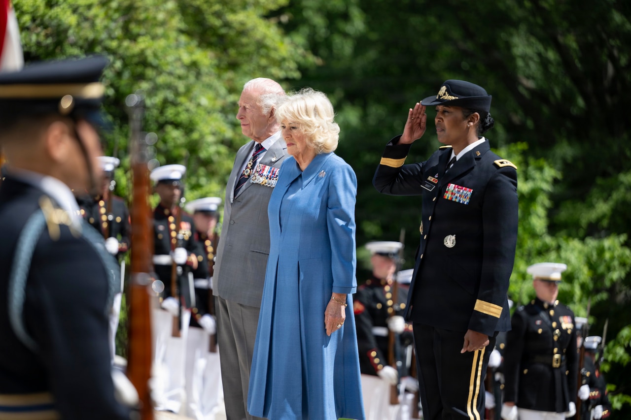 A man wearing a gray suit and a woman in a blue dress stand at attention while a woman in military dress uniform salutes. Rows of other people wearing military dress uniforms stand around them.