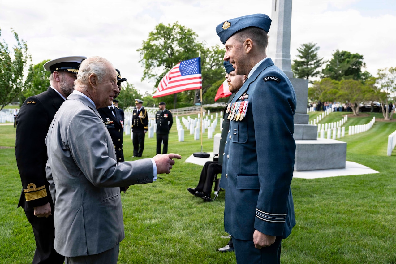 A man, dressed in a suit, smiles at two men wearing dress military uniforms, one is wearing a foreign military uniform. A cemetery memorial, graves and an American flag are in the background.