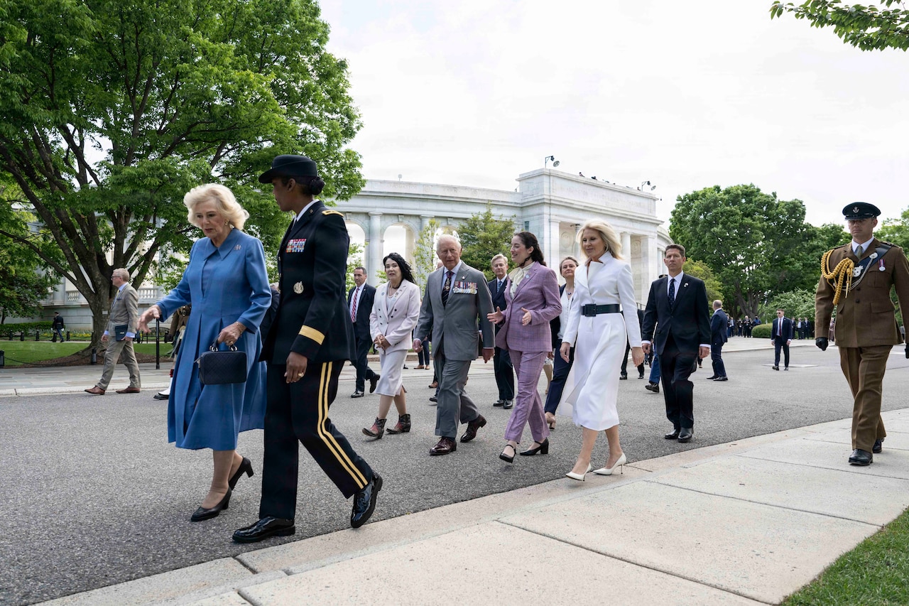 A group of people walk on a paved road. Some people are wearing military dress uniforms while some men are wearing suits and women are wearing dresses. A marble building is in the background.
