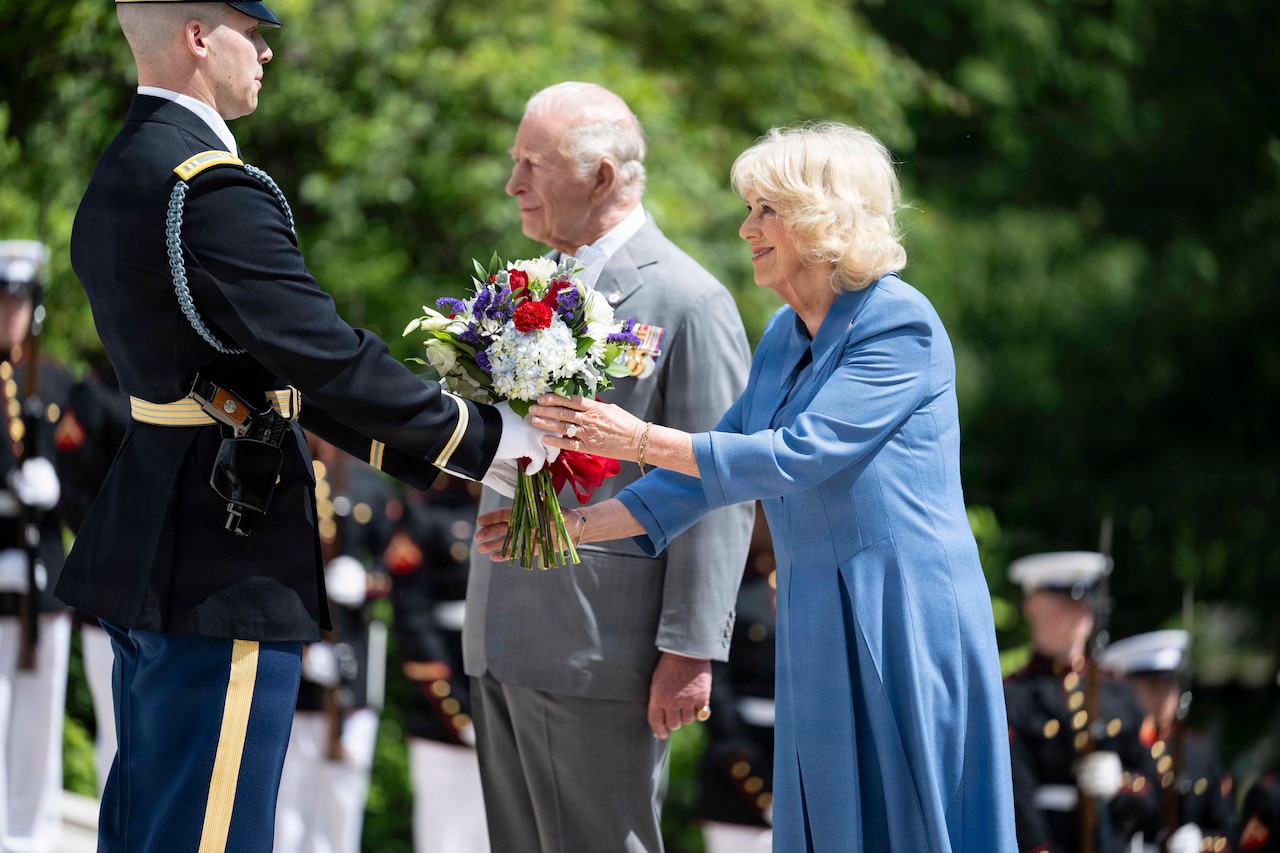 A man wearing a dress military uniform hands a woman in a blue dress a bouquet of flowers. A man in a gray suit is standing next to her.