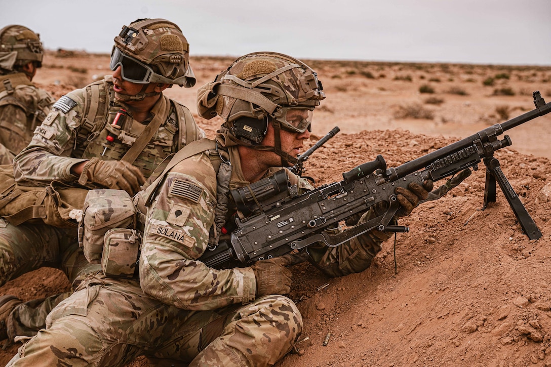 Three soldiers in tactical gear lie in a dirt trench as one aims a weapon during the day.