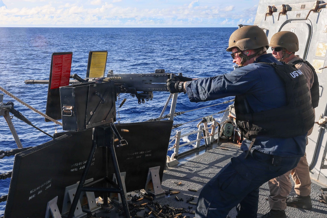 A sailor fires a weapon off the side of a ship at sea as fellow service member watches.