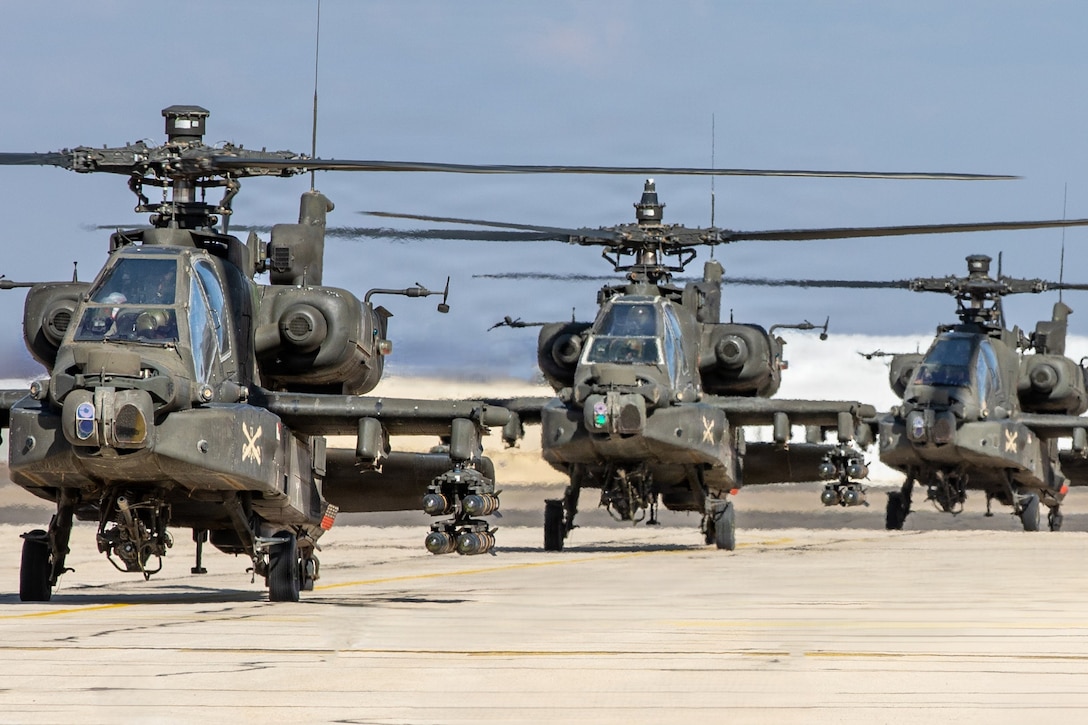 Three helicopters move in formation on a tarmac under a gray sky.
