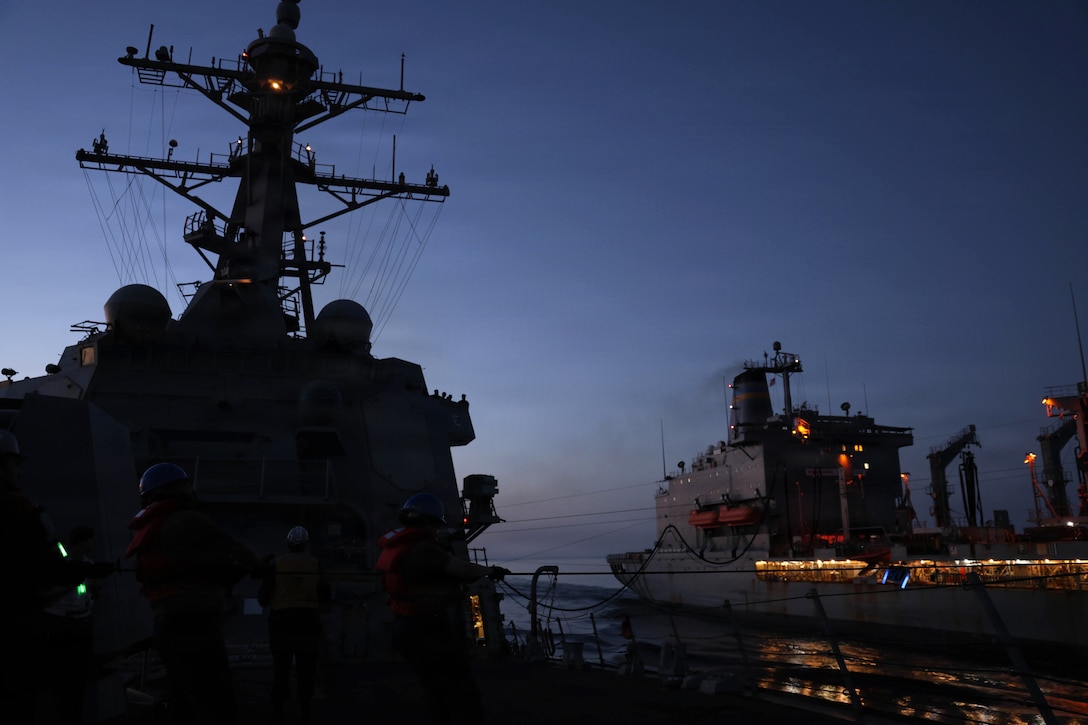 Two ships connected by lines sail next to each other in the dark illuminated by a ship's lights.