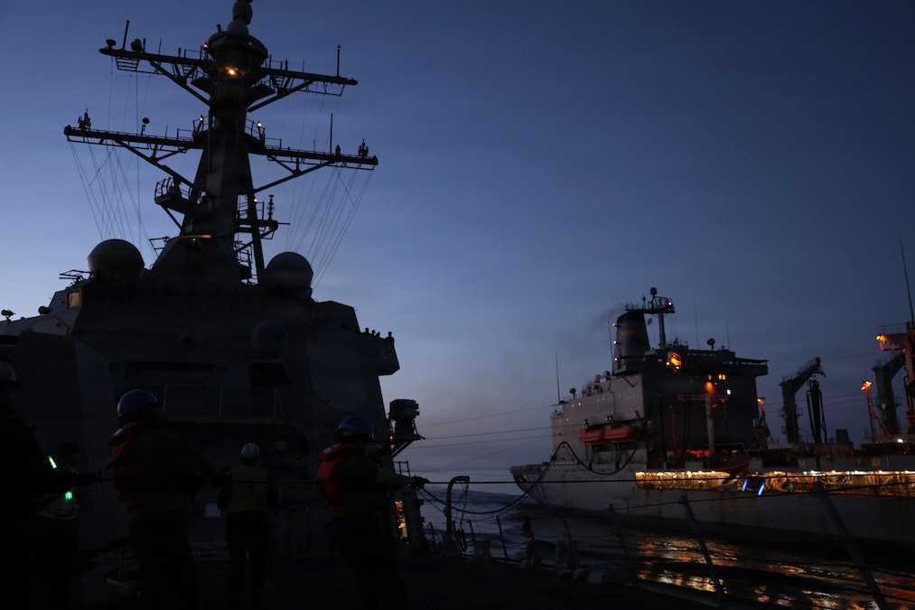 Two ships connected by lines sail next to each other in the dark illuminated by a ship's lights.