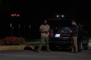 Two men wearing casual attire stand next to a car outside at night, holding remote controls and operating drones.
