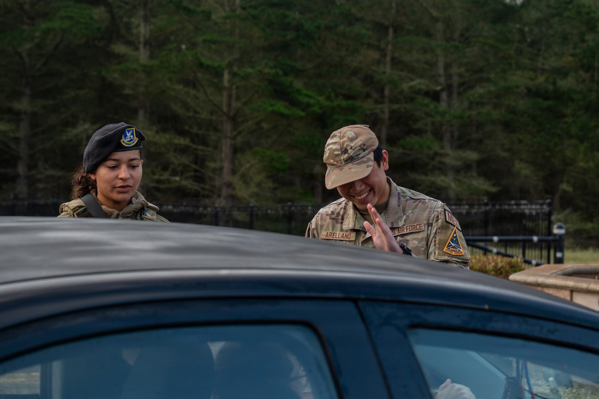 Two people stand next to each other a greet a person in a car.