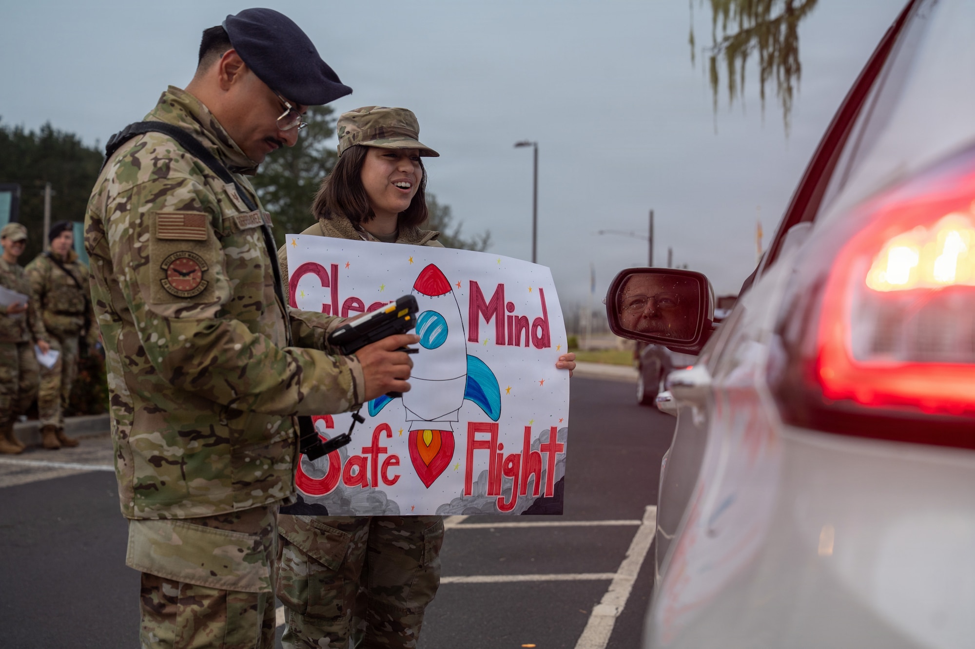 Two people stand next to a car, one of the people hold a sign.