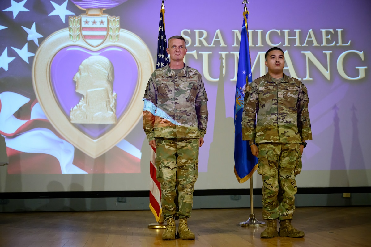 Two men in camouflage stand at attention on a stage.