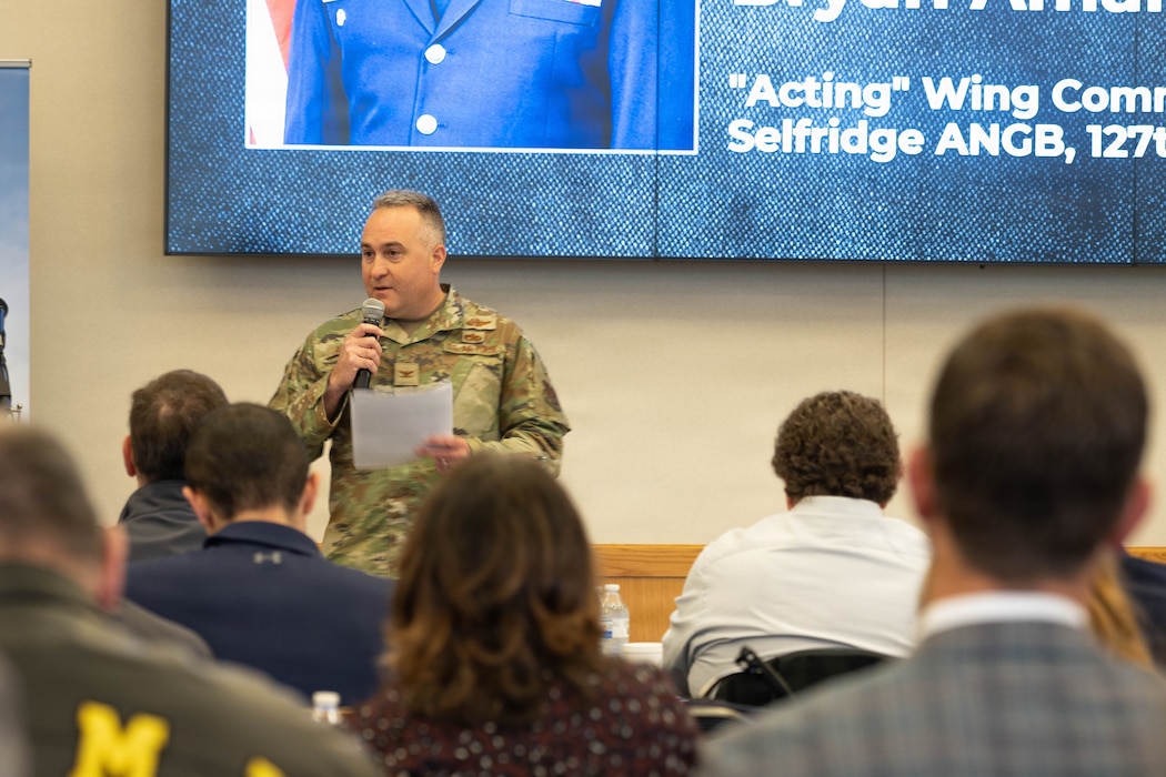 Air Force officer in camo green uniform address audience of civilians while holding a piece of paper and a microphone.