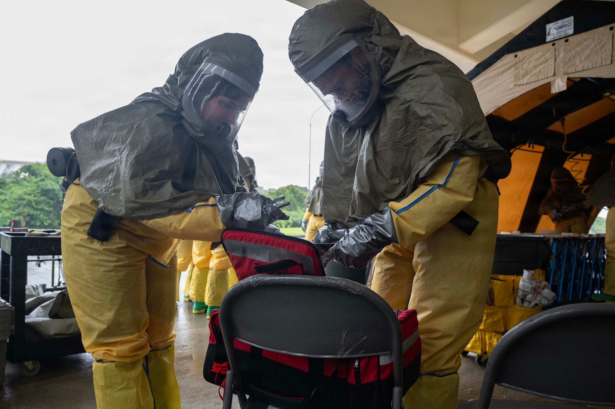 Airmen in personal protective suits in a tent grab supplies from a red medical bag.