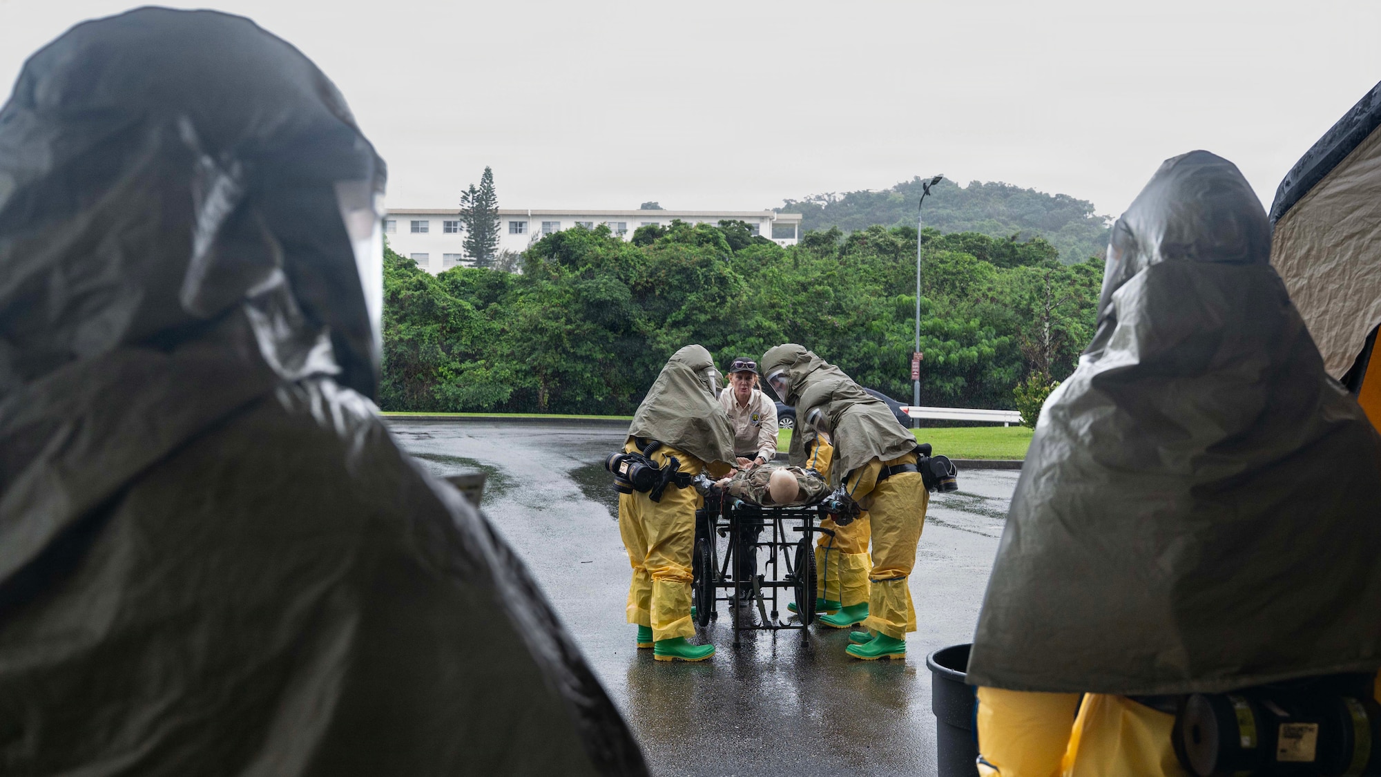 Airmen in personal protective suits stand around a mannequin on a stretcher in the rain.