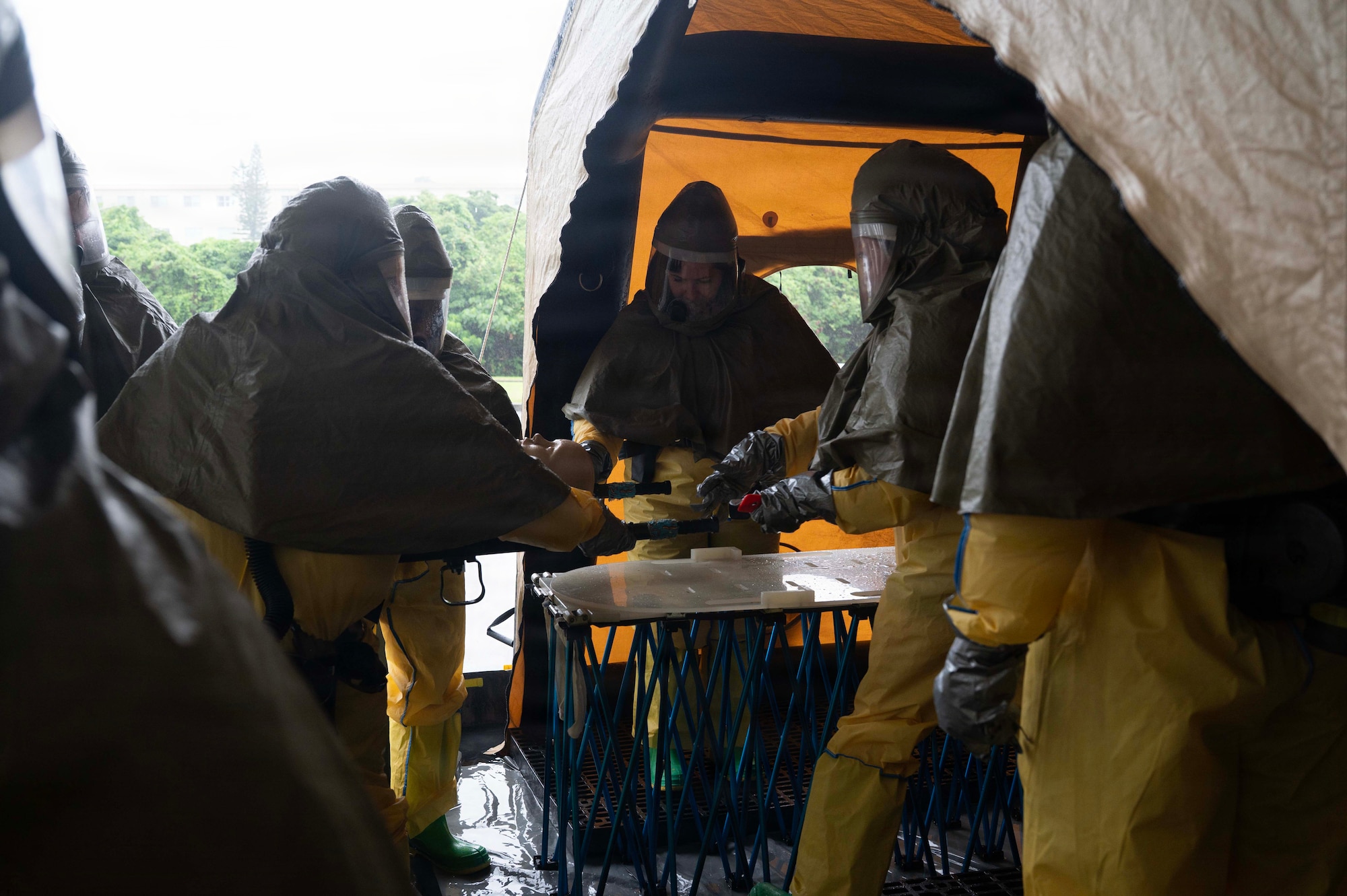 Airmen in personal protective suits transfer off a mannequin to others in a tent.