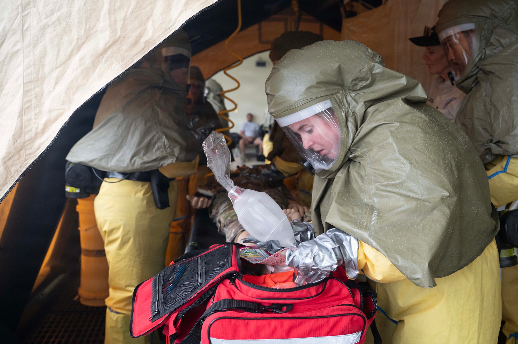 A person in personal protective suits in a tent grabs medical supplies out of a bag while others in the background work in a tent.