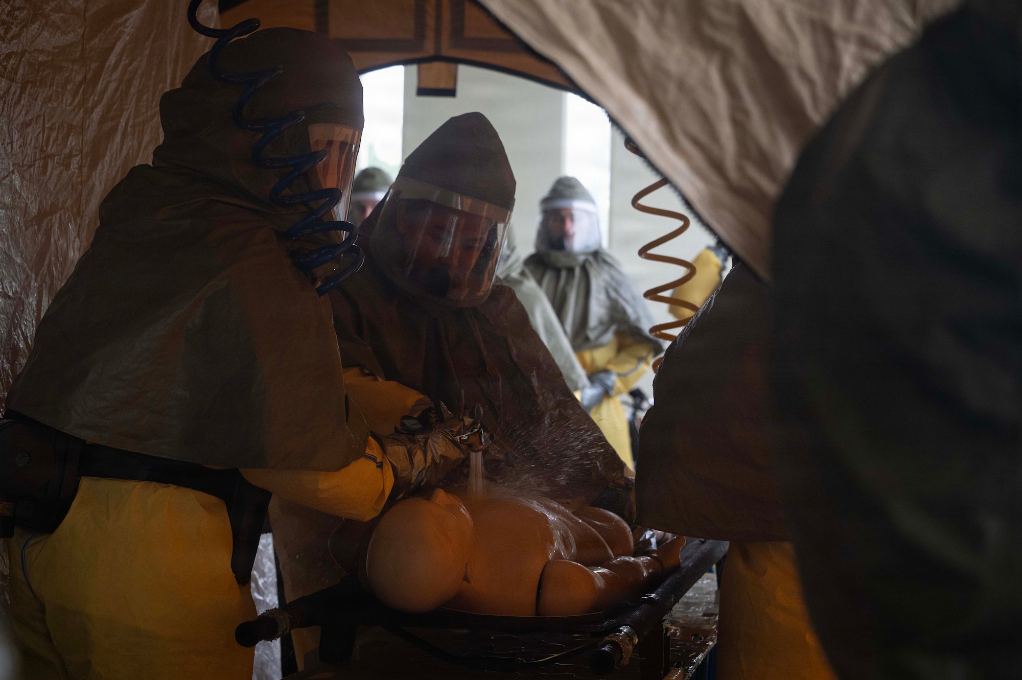 People wash down a mannequin with a water spray while in personal protective suits in a tent.