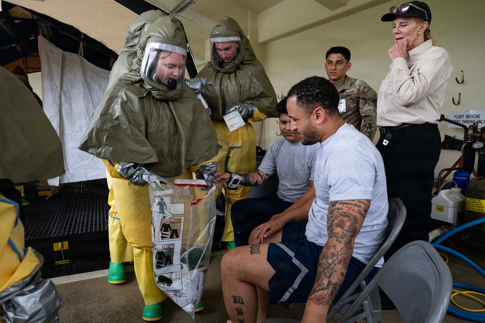 A person holds a bag while in a personal protective suit with someone sitting down and people in the background.